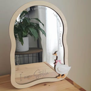 May include: An arched wooden mirror with a wavy frame featuring a white goose with a pink bow attached to the bottom right. The mirror surface is etched with the phrases I am smart, I am worthy, I am unique, I am loved, I am kind, and I am Stella.