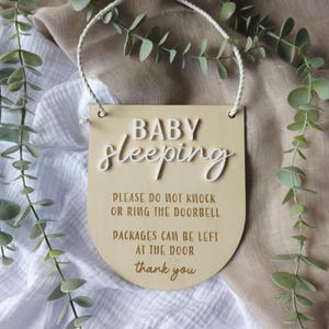 May include: A wooden sign with the words "BABY sleeping" in white script. The sign also includes the text "PLEASE DO NOT KNOCK OR RING THE DOORBELL PACKAGES CAN BE LEFT AT THE DOOR thank you". A white rope is attached for hanging.