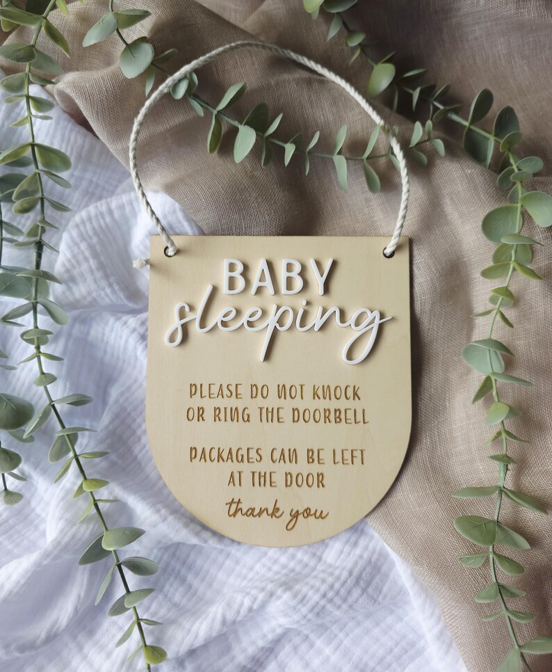 May include: A wooden sign with the words "BABY sleeping" in white script. The sign also includes the text "PLEASE DO NOT KNOCK OR RING THE DOORBELL PACKAGES CAN BE LEFT AT THE DOOR thank you". A white rope is attached for hanging.
