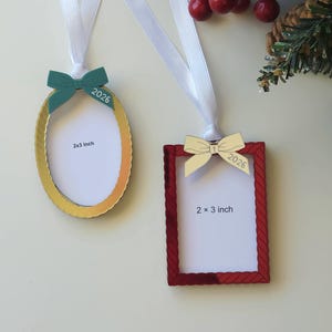 May include: Two hanging picture frame ornaments with white ribbons. The oval frame is gold with a teal bow and the text 2026. The rectangular frame is red with a cream bow and the text 2026. Both frames feature white inserts labelled 2x3 inch.