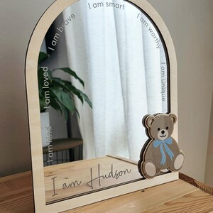 May include: An arched mirror with a light wood frame featuring a layered wooden teddy bear with a blue bow on the bottom right. The mirror surface is etched with the phrases I am smart, I am worthy, I am unique, I am brave, I am loved, I am kind, and I am Hudson.