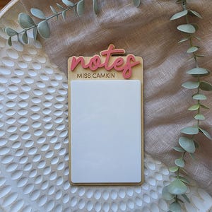May include: A wooden notepad with the word "notes" in pink script at the top, and the name "MISS CAMKIN" below. The notepad has a white rectangular space for writing. The notepad is on a white textured surface with green leafy sprigs.