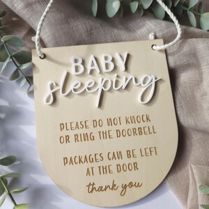 May include: Wooden sign with the words "BABY sleeping" in white raised letters. The sign also includes the text "PLEASE DO NOT KNOCK OR RING THE DOORBELL PACKAGES CAN BE LEFT AT THE DOOR thank you". A white string is attached for hanging.