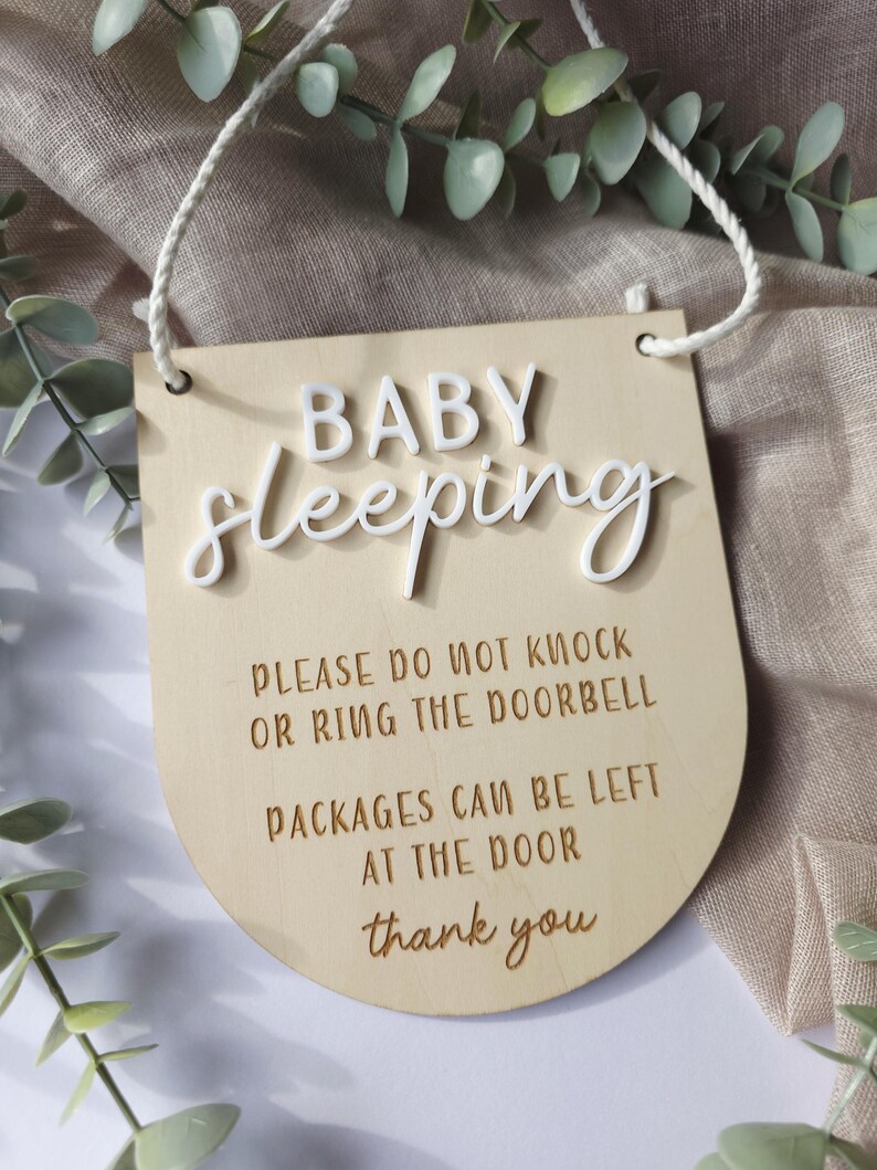 May include: Wooden sign with the words "BABY sleeping" in white raised letters. The sign also includes the text "PLEASE DO NOT KNOCK OR RING THE DOORBELL PACKAGES CAN BE LEFT AT THE DOOR thank you". A white string is attached for hanging.