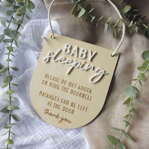May include: A wooden sign with the words "BABY Sleeping" in white script. The sign also includes the phrases "PLEASE DO NOT KNOCK OR RING THE DOORBELL" and "PACKAGES CAN BE LEFT AT THE DOOR". A white rope is attached for hanging.