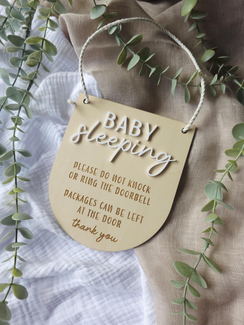 May include: A wooden sign with the words "BABY Sleeping" in white script. The sign also includes the phrases "PLEASE DO NOT KNOCK OR RING THE DOORBELL" and "PACKAGES CAN BE LEFT AT THE DOOR". A white rope is attached for hanging.