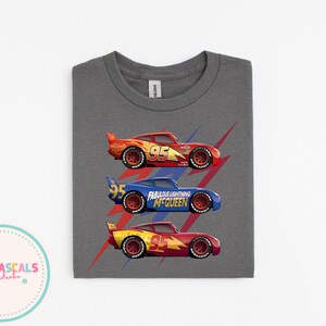 Puede incluir: Una camiseta gris con tres coches de carreras de dibujos animados en rojo, azul y amarillo. Cada coche tiene el n&uacute;mero 95. El coche azul tiene el texto "Fabulous Lightning McQueen". Rayos rojos detr&aacute;s de los coches.