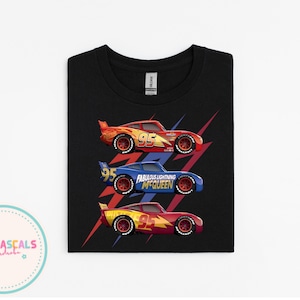 Puede incluir: Camiseta negra con tres coches de carreras de dibujos animados en rojo, azul y amarillo. El coche azul tiene el texto "Fabulous Lightning McQueen". La camiseta est&aacute; doblada y lista para usar.
