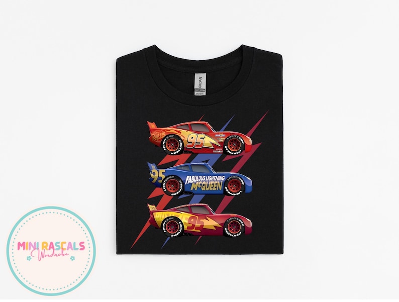 Puede incluir: Camiseta negra con tres coches de carreras de dibujos animados en rojo, azul y amarillo. El coche azul tiene el texto "Fabulous Lightning McQueen". La camiseta est&aacute; doblada y lista para usar.