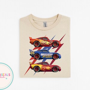 Puede incluir: Una camiseta beige con tres coches de carreras rojos, azules y amarillos, cada uno con el n&uacute;mero 95. Los coches est&aacute;n sobre un dise&ntilde;o de rayo rojo y azul. El texto "Fabulous Lightning McQueen" es visible.