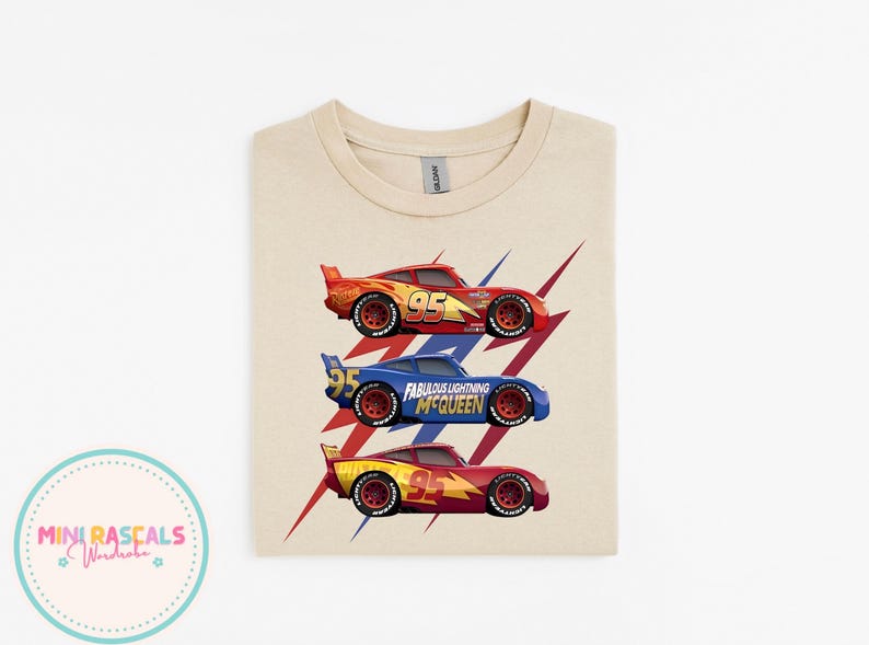 Puede incluir: Una camiseta beige con tres coches de carreras rojos, azules y amarillos, cada uno con el n&uacute;mero 95. Los coches est&aacute;n sobre un dise&ntilde;o de rayo rojo y azul. El texto "Fabulous Lightning McQueen" es visible.