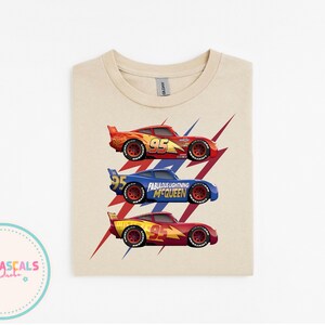 Puede incluir: Una camiseta beige con tres coches de carreras de dibujos animados en rojo, azul y amarillo. Cada coche tiene el n&uacute;mero 95. El coche azul tiene el texto "Fabulous Lightning McQueen". Gr&aacute;ficos de rayos rojos y azules detr&aacute;s de los coches.