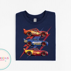 Puede incluir: Camiseta azul marino con tres coches de carreras rojos, azules y amarillos. Cada coche tiene el n&uacute;mero 95. El coche azul tiene el texto "Fabulous Lightning McQueen". Rayos rojos detr&aacute;s de cada coche.