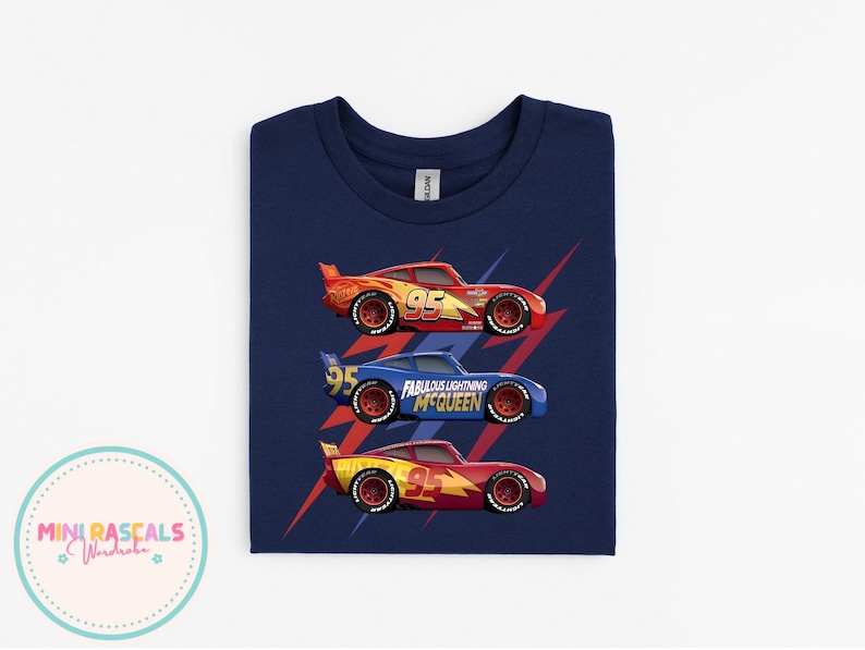 Puede incluir: Camiseta azul marino con tres coches de carreras rojos, azules y amarillos. Cada coche tiene el n&uacute;mero 95. El coche azul tiene el texto "Fabulous Lightning McQueen". Rayos rojos detr&aacute;s de cada coche.