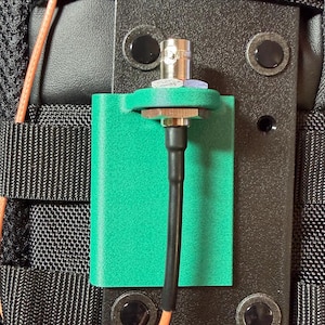 Puede incluir: Primer plano de una placa de montaje negra con un soporte verde que sujeta un conector plateado y un cable negro. La placa está fijada a una tela negra con cableado naranja visible.
