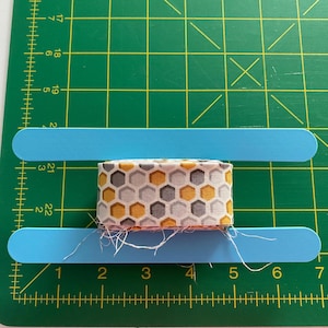 May include: A fabric ribbon with a honeycomb pattern in yellow, gray, and white is positioned between two blue craft sticks. The ribbon is on a green cutting mat with measurement markings.