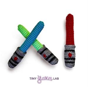 May include: Crocheted toy lightsabers in green, blue, and red. The handles are gray with black stripes and a red button. The text "Tiny Yarn Lab" is at the bottom.