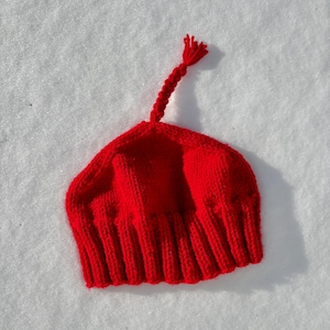 May include: A bright red knitted hat with a braided tassel. The hat is made of a thick yarn and has a ribbed brim. The hat is lying on a white, snowy surface.