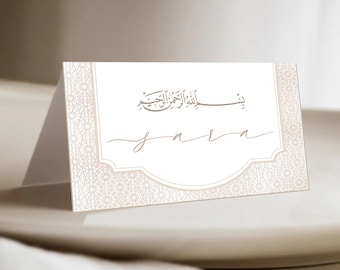 May include: A white place card with a brown border and intricate design. The card features the Arabic phrase "Bismillah al-Rahman al-Rahim" and the name "Sara" in a cursive script.