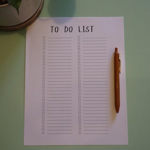 May include: A white To Do List with a brown pen on a light green surface. The list has two columns of checkboxes and horizontal lines for writing. The words "TO DO LIST" are printed at the top in a handwritten style font.