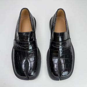 Peut inclure: Paire de mocassins noirs en cuir avec un design à bout fendu. Les chaussures ont une texture brillante, semblable à celle d'un crocodile, et une semelle épaisse et arrondie. L'intérieur est de couleur beige clair.