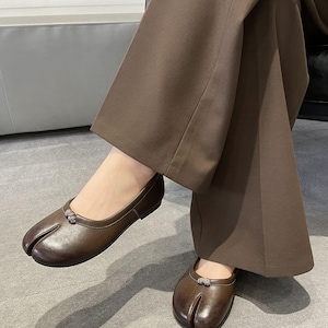 May include: Brown leather split-toe ballet flats. The shoes have a small decorative embellishment on the top and are worn with brown wide-leg pants. The shoes have a unique design with a split between the big toe and the other toes.