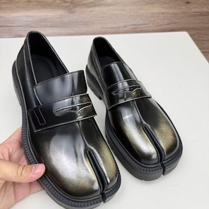 Op de afbeelding: Een paar zwarte leren split-toe loafers met een glanzende afwerking. De schoenen hebben een uniek ontwerp met een apart gedeelte voor de grote teen en een dikke, geribbelde zool. De loafers worden op een wit oppervlak weergegeven.