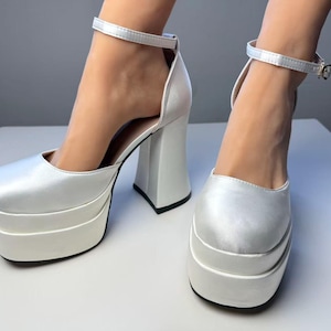 May include: White platform heels with a square toe and ankle strap. The chunky heel and platform sole are white, with a black sole. The shoes have a satin finish and a classic design.