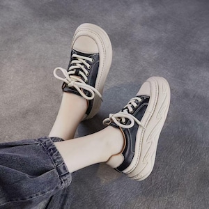 以下が含まれることがあります： 白の靴紐が付いた、クリーム色と黒色のスニーカー。靴は厚く波状のソールを備えています。スニーカーは紐で締められています。ソールの厚さは約3cmです。