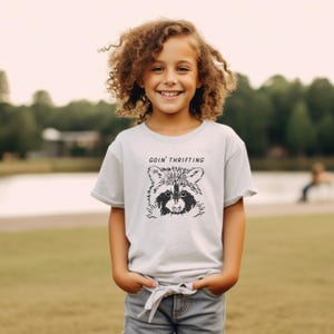 May include: A light gray t-shirt featuring a black and white raccoon graphic with the text "GOIN' THRIFTING". The shirt is worn by a young person with curly hair and blue jeans. The background is a green field and trees.