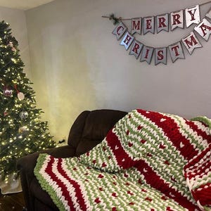 May include: A red, white and green striped knitted throw blanket with a heart pattern on a brown sofa. A Christmas tree with lights and baubles is in the background. A banner with the text "Merry Christmas" is hanging on the wall.
