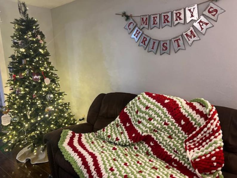 May include: A red, white and green striped knitted throw blanket with a heart pattern on a brown sofa. A Christmas tree with lights and baubles is in the background. A banner with the text "Merry Christmas" is hanging on the wall.