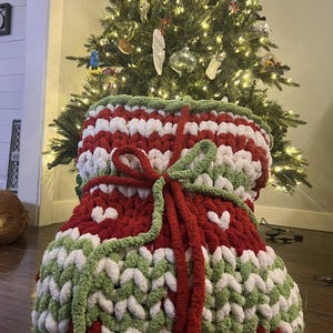 May include: A large, knitted Christmas stocking in red, white and green with a white heart pattern. The stocking is tied with a red ribbon and is sitting in front of a Christmas tree.