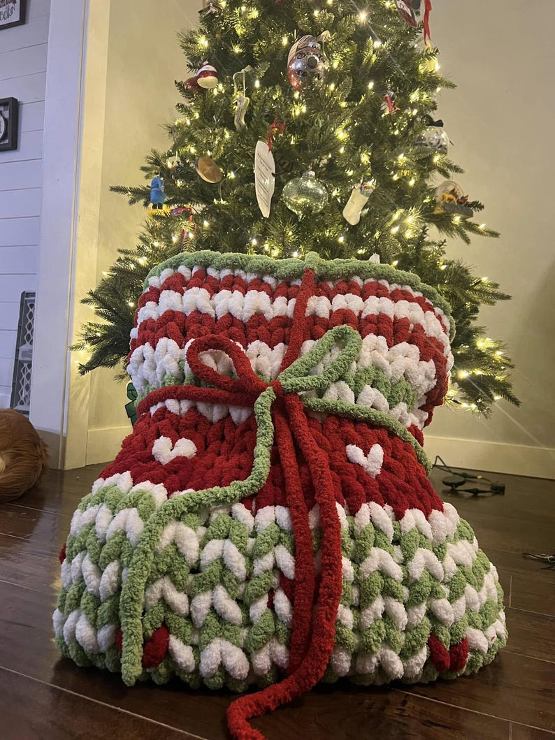 May include: A large, knitted Christmas stocking in red, white and green with a white heart pattern. The stocking is tied with a red ribbon and is sitting in front of a Christmas tree.