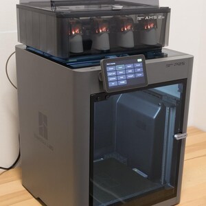 May include: A gray 3D printer with a clear door and a touch screen interface. The printer has a black top section with multiple compartments. The brand name "Bambu Lab" is visible on the front.