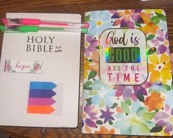 Può includere: Una Sacra Bibbia bianca con il testo "Holy Bible KJV" e un adesivo floreale "hope". Accanto, un quaderno floreale con la scritta "God is Good All the Time". Penne rosa e verdi sopra.