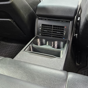 Könnte beinhalten: Innenansicht eines Autoinnenraums mit schwarzer Mittelkonsole, Armlehne, Lüftungsschlitzen und Getränkehaltern. Die Konsole ist zwischen den Vordersitzen integriert und betont Funktionalität und Design.