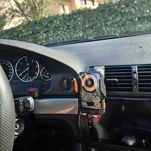 May include: A black car dashboard with a phone holder attached to the air vent. The phone holder has a circuit board design on the back and an orange accent. The car's speedometer and other gauges are visible.