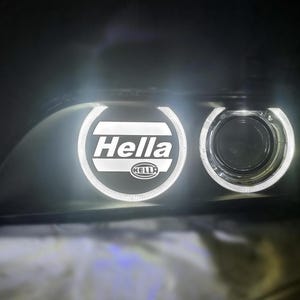 May include: A close-up of a car headlight with a white ring around the projector lens. The lens has a Hella logo in the center.