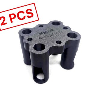May include: A dark gray, four-legged mechanical component with a flat top surface featuring multiple circular holes. The top is engraved with the text M57N2 Rev3_202410. A red rectangular frame in the corner contains the text 2 PCS.