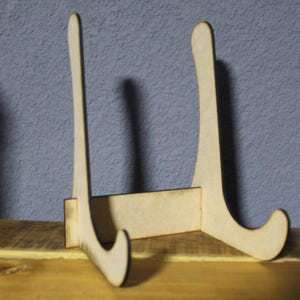 Puede incluir: Un soporte de exhibición de madera de color marrón claro. El soporte tiene dos soportes curvos y una base rectangular. Diseñado para exhibir platos u objetos decorativos. El soporte está sobre una superficie de madera.