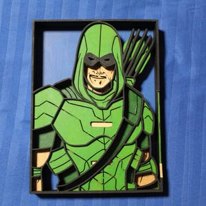 May include: A layered wooden art piece featuring a superhero in a green costume with a hood and mask, holding a bow and arrows. The artwork is framed in black and set against a blue background.