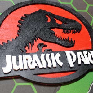 May include: A wooden Jurassic Park logo featuring a black dinosaur silhouette on a red background, with the words "JURASSIC PARK" in white letters. The logo is set against a gray and green hexagonal pattern.