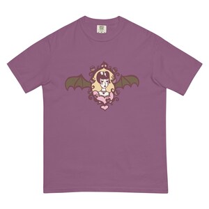 May include: Purple t-shirt with a graphic of a girl with wings and a heart. The graphic is in shades of pink, brown, and white.