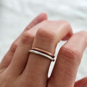 May include: Two minimalist rings, one silver and one copper, stacked on a finger. The rings have a simple, band design. The background is a soft, white fabric.