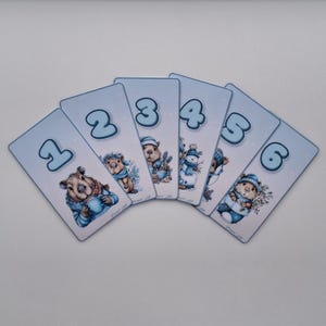 Può includere: Sei carte azzurre con i numeri da 1 a 6. Ogni carta presenta una diversa illustrazione di animali a tema invernale, tra cui una marmotta, un riccio e un pupazzo di neve. Le carte sono disposte a ventaglio.