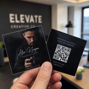 May include: Two black square business cards are held in a hand. One card features a portrait and the text "Alex Morgan Photography." The other card has a QR code and the text "Capturing moments that last forever. Scan to view portfolio."
