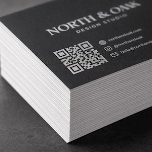 May include: A stack of rectangular business cards with a matte black finish and white text. The cards feature the text NORTH & OAK DESIGN STUDIO, a square QR code, and contact information including a website, Instagram handle, and email address.