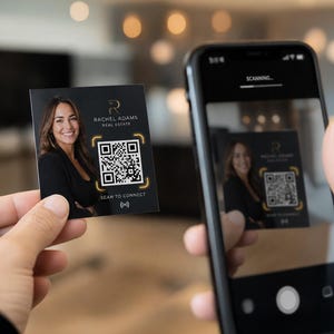 May include: A business card with the text "RACHEL ADAMS REAL ESTATE" and a QR code is held in front of a smartphone. The phone screen displays a scanning interface. The card also has the text "SCAN TO CONNECT".