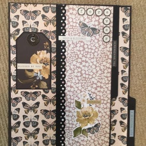 May include: A decorative scrapbook with a butterfly and floral design. The cover features a tag that says "Blessed by You". The scrapbook has the words "forever" and "remembering you" on it. The color scheme includes shades of brown, beige, and black.